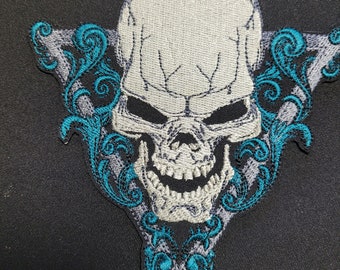 Puede incluir: Una calavera blanca con una sonrisa de dientes está rodeada de bordados florales de color azul turquesa y gris. La calavera está centrada en un diseño triangular.