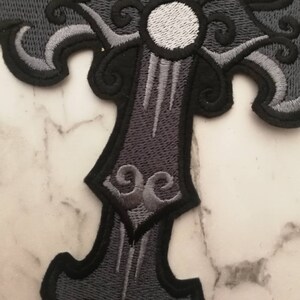 May include: A black and gray embroidered iron-on patch featuring a stylized cross with a white circle in the center. The cross has intricate details and a faded look.