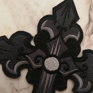 May include: A black and gray embroidered cross patch with a decorative design. The cross has a white center circle.