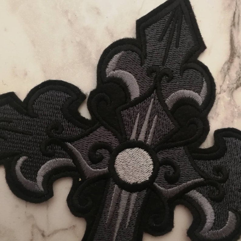 May include: A black and gray embroidered cross patch with a decorative design. The cross has a white center circle.