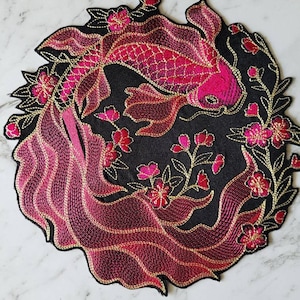 Può includere: Una toppa ricamata con un pesce koi che nuota attraverso un cerchio di fiori e foglie rosa su uno sfondo nero. Il pesce è rosa e dorato.