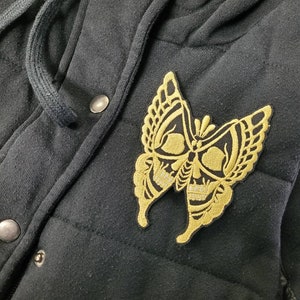 May include: Black hooded jacket with a gold embroidered butterfly design. The butterfly has a skull-like pattern.
