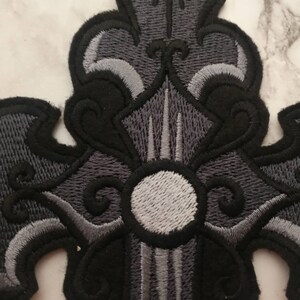 May include: Black and gray embroidered cross patch with a white circle in the center.