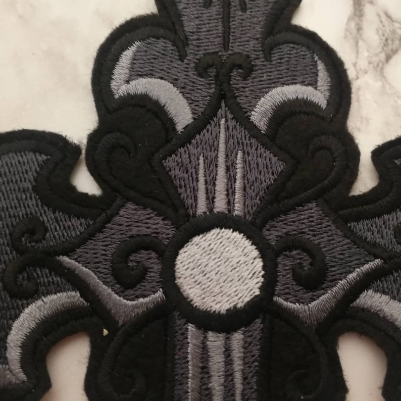 May include: Black and gray embroidered cross patch with a white circle in the center.