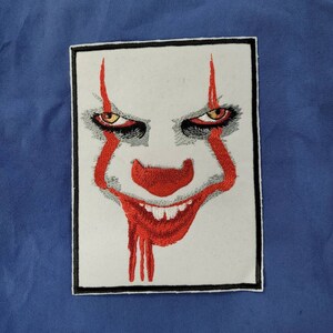 Może przedstawiać: Naszywka haftowana przedstawiająca zbliżenie twarzy klauna z czerwono-białym makijażem, czerwonym nosem i złowrogim uśmiechem. Oczy klauna są żółte i czarne.