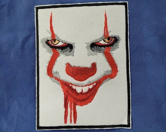 Puede incluir: Parche bordado con un primer plano de la cara de un payaso con maquillaje rojo y blanco, una nariz roja y una sonrisa siniestra. Los ojos del payaso son amarillos y negros.
