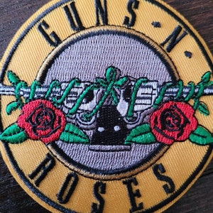 May include: An embroidered patch featuring the words "Guns N' Roses" in a circle around a skull and crossbones with roses, in yellow and black.