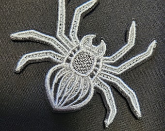 Puede incluir: Un parche de araña bordado en blanco con patas detalladas y un cuerpo texturizado.