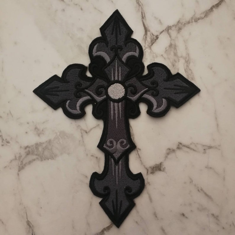 May include: A black and gray embroidered cross with intricate details. The cross has a white center circle and is outlined in black.