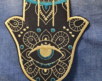 Puede incluir: Un parche bordado en negro y dorado con un diseño de mano de hamsa y un ojo en el centro. El parche tiene un borde azul y dorado.