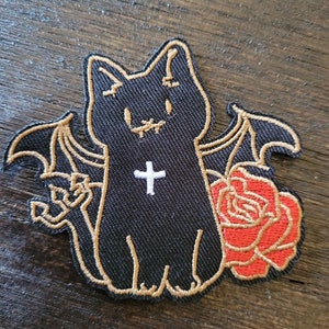 Może przedstawiać: Czarna haftowana naszywka do prasowania przedstawiająca kota z skrzydłami nietoperza, krzyżem na piersi i czerwoną różą.