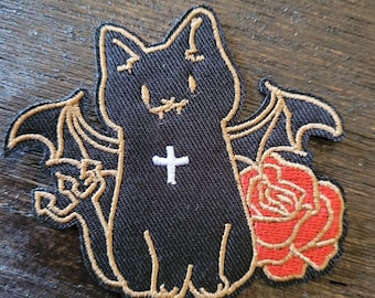 Puede incluir: Parche de hierro negro bordado de un gato con alas de murciélago, una cruz en el pecho y una rosa roja.