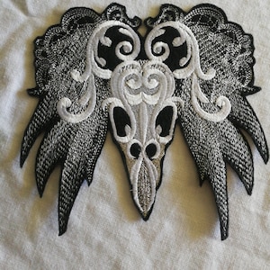 May include: Black and white embroidered patch featuring a skull design with intricate floral details. The skull has a pointed chin and two large eye sockets.