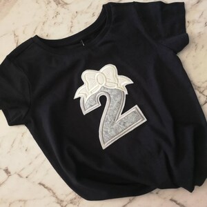 May include: A black t-shirt with a silver number two with a white bow on top. The number is a patch sewn onto the shirt.