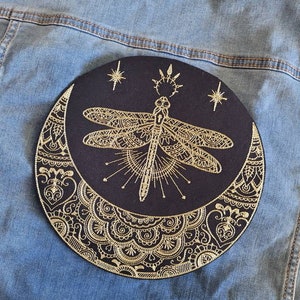 May include: A black fabric patch with a gold embroidered design of a dragonfly, crescent moon, and stars. The design is intricate and detailed, with a mandala-like pattern around the moon.