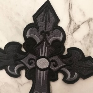 May include: A black and gray embroidered cross patch with intricate details. The cross is a traditional design with a fleur-de-lis at the top and bottom.