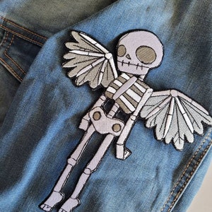 May include: Embroidered patch featuring a skeleton with angel wings, in shades of gray and white. The patch is sewn onto a denim jacket, showcasing the design's details and texture. Ideal for adding a unique touch to clothing or accessories.