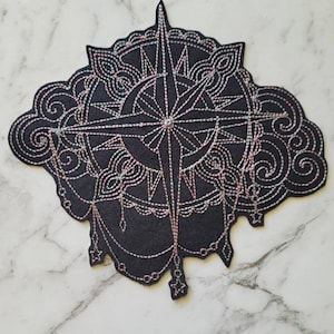 May include: A black fabric patch with a white and pink embroidered compass design. The compass is surrounded by a decorative mandala-like pattern.