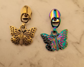 Puede incluir: Dos tiradores de cremallera con dijes de mariposa. Un tirador es de oro antiguo con un dije de mariposa dorado. El otro tirador es iridiscente con un dije de mariposa arcoíris.