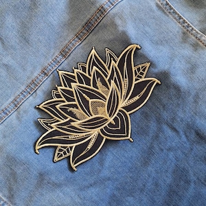 Może przedstawiać: Naszywka z haftowanym kwiatem lotosu w kolorze czarnym i złotym. Naszywka znajduje się na niebieskim denimie.