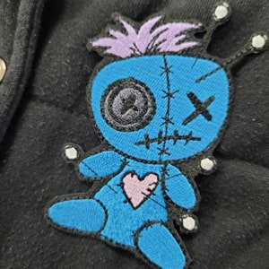 May include: A blue and purple embroidered patch of a voodoo doll with a pink heart on its chest. The doll has stitches around its eyes and mouth, and a cross on its forehead.