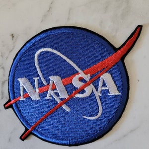 May include: Embroidered patch featuring the NASA logo. The patch has a blue circular background with the word "NASA" in white, and a red swoosh. A white arc curves through the centre of the logo.