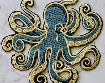Puede incluir: Un pulpo azul turquesa con tentáculos bordados en oro. El pulpo está contorneado en negro y tiene una superficie texturizada.