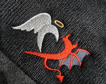 Puede incluir: Gorro negro con un diseño bordado de un ángel y un diablo. El ángel tiene alas blancas y un halo dorado, mientras que el diablo tiene cuernos rojos y una cola.