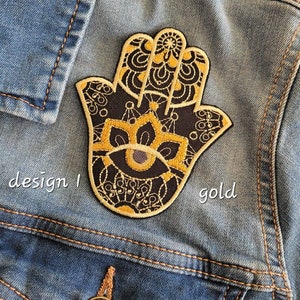 Può includere: Una toppa ricamata in oro a forma di mano di Hamsa con un design mandala in bianco e nero. La toppa è su una giacca di jeans blu.