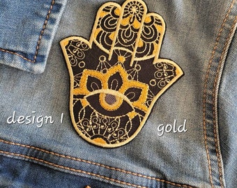 Puede incluir: Un parche bordado en dorado de una mano de hamsa con un diseño de mandala en blanco y negro. El parche está en una chaqueta vaquera azul.