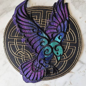May include: An embroidered patch featuring a purple and blue bird with outstretched wings, set against a black background with a gold Celtic knot design.