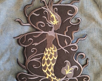 Puede incluir: Parche bordado en negro y dorado de una sirena con el pelo suelto y una cola con escamas. La sirena está rodeada de enredaderas en espiral.