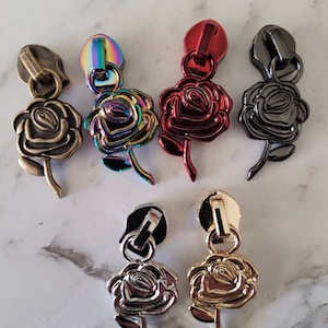 May include: Six zipper pulls with rose-shaped charms in various colours, including gold, silver, black, red, and iridescent.