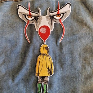 Może przedstawiać: Naszywka haftowana przedstawiająca twarz klauna z czerwonym i białym makijażem, trzymającego czerwony balon. Postać w żółtym płaszczu przeciwdeszczowym i zielonych butach idzie w kierunku klauna.