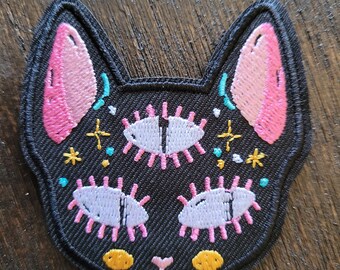 Puede incluir: Parche de gato negro bordado con orejas rosas, ojos blancos y detalles amarillos. El gato tiene un tercer ojo en el centro de la frente.