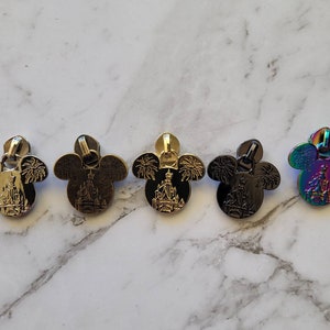 May include: Five zipper pulls in different finishes, each featuring a Mickey Mouse head silhouette with a silhouette of Cinderella's castle and fireworks inside.