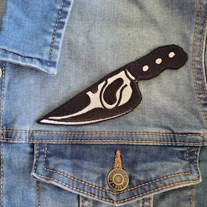 May include: A black and white embroidered patch of a knife on a blue denim jacket.