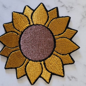 Könnte beinhalten: Ein gestickter Sonnenblumen-Patch in Gelb mit brauner Mitte. Die Sonnenblume hat einen schwarzen Umriss und ist perfekt, um Ihren Projekten einen Hauch von Natur zu verleihen.
