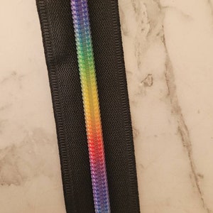 May include: A black zipper with a rainbow colored zipper pull. The zipper is on a white marble background.