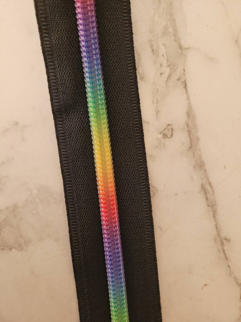 May include: A black zipper with a rainbow colored zipper pull. The zipper is on a white marble background.