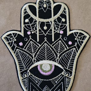 May include: Black Hamsa hand patch with intricate white embroidery. The design includes an eye, leaf patterns, and small pink dots. The patch has a beige border and is likely for clothing or accessories.