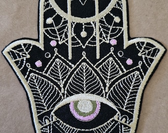Puede incluir: Parche negro con forma de mano de Hamsa con intrincado bordado blanco. El diseño incluye un ojo, patrones de hojas y pequeños puntos rosas. El parche tiene un borde beige, para ropa o accesorios.