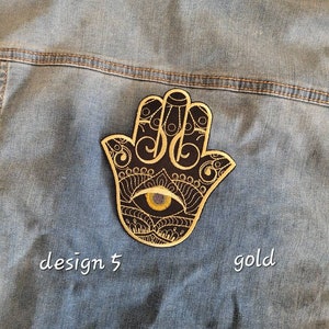 Può includere: Patch ricamato in oro a forma di mano di Hamsa su sfondo nero con un occhio giallo al centro. Il patch è su una giacca di jeans blu. Il testo "design 5" e "gold" è scritto sotto il patch.