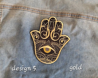 Puede incluir: Parche bordado en oro con forma de mano de Hamsa sobre fondo negro con un ojo amarillo en el centro. El parche está en una chaqueta vaquera azul. El texto "design 5" y "gold" está escrito debajo del parche.