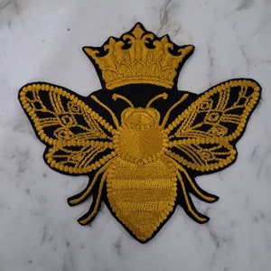 May include: A gold embroidered bee with wings and a crown on a black background. The bee is a popular motif in fashion and design.