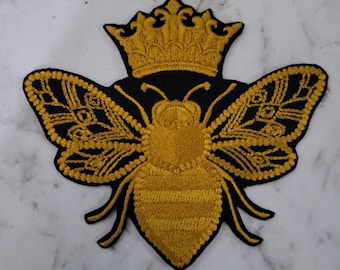 Puede incluir: Una abeja bordada en oro con alas y una corona sobre un fondo negro. La abeja es un motivo popular en la moda y el diseño.