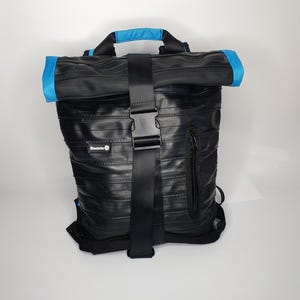 May include: A black roll-top backpack constructed from recycled rubber inner tubes, featuring bright blue accents on the top handle and roll-top edges. A central black nylon strap with a plastic buckle secures the closure, and a small label reads Becicle.