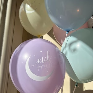 May include: A set of four balloons in various pastel colours, including light blue, lavender, light pink, and cream. The lavender balloon has the text "eid mubarak" written in white cursive with a crescent moon and stars.