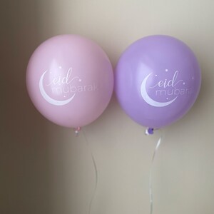 May include: Two balloons, one pink and one purple, with the text "eid mubarak" in white lettering. The balloons are tied with white strings.