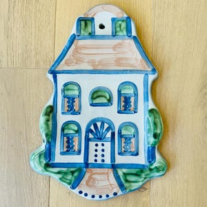 Puede incluir: Un adorno de pared de cerámica con forma de casa, con base blanca, ribetes azules y tejado marrón. La casa presenta ventanas arqueadas con detalles azules y naranjas, una puerta azul y árboles verdes a ambos lados.